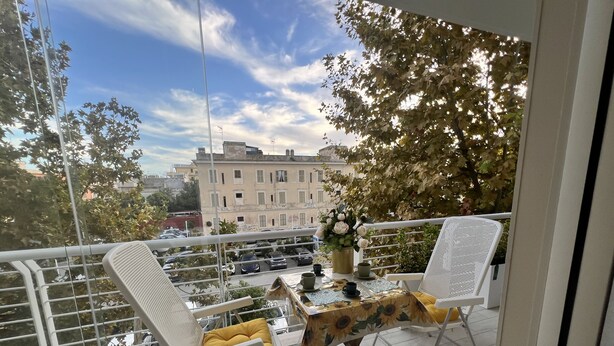 Een balkon met twee witte stoelen, een tafel, een bloempot en uitzicht op gebouwen en geparkeerde auto's.