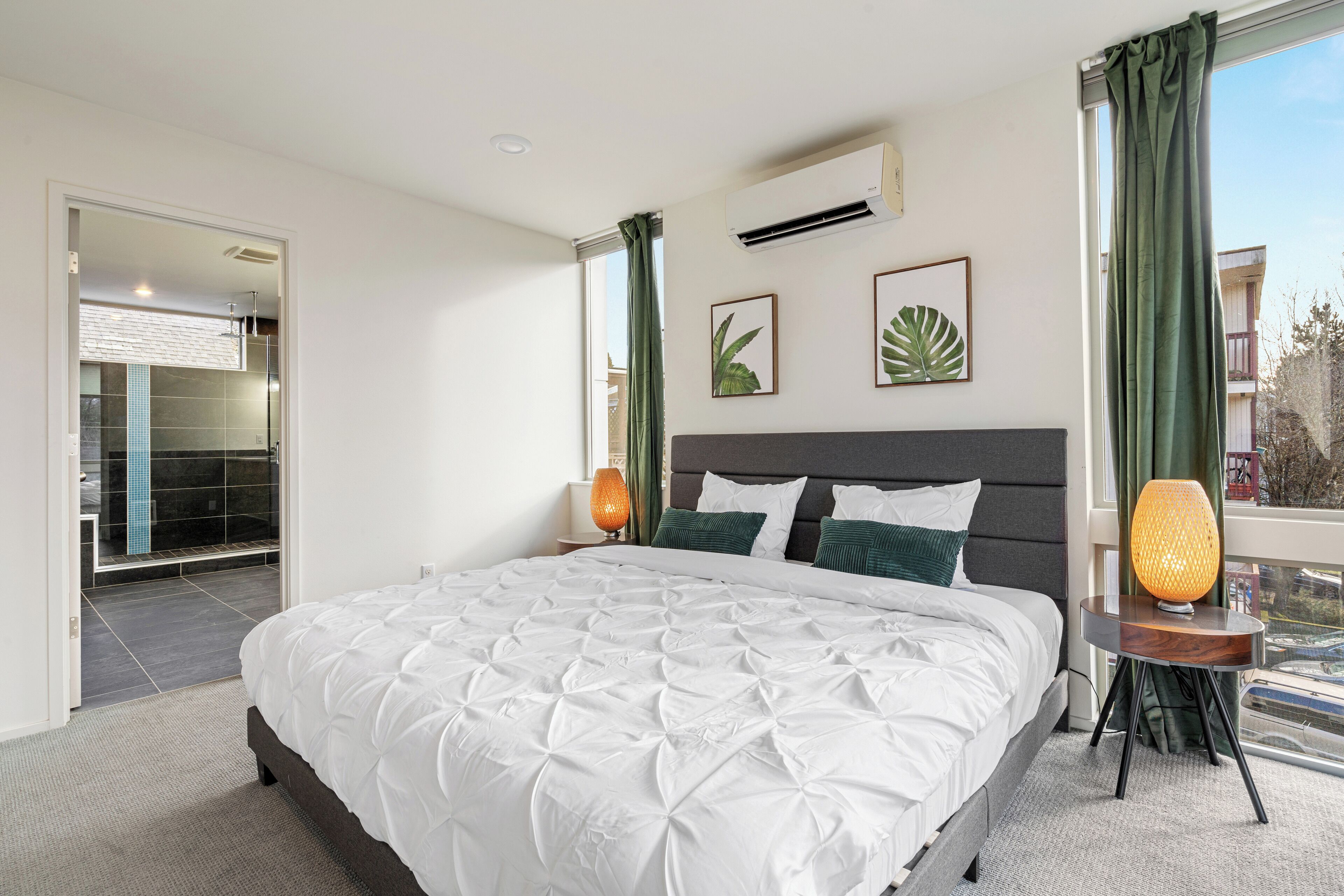 A modern hotel room with a large bed, a nightstand with a lamp, and a view of the outside through a window.