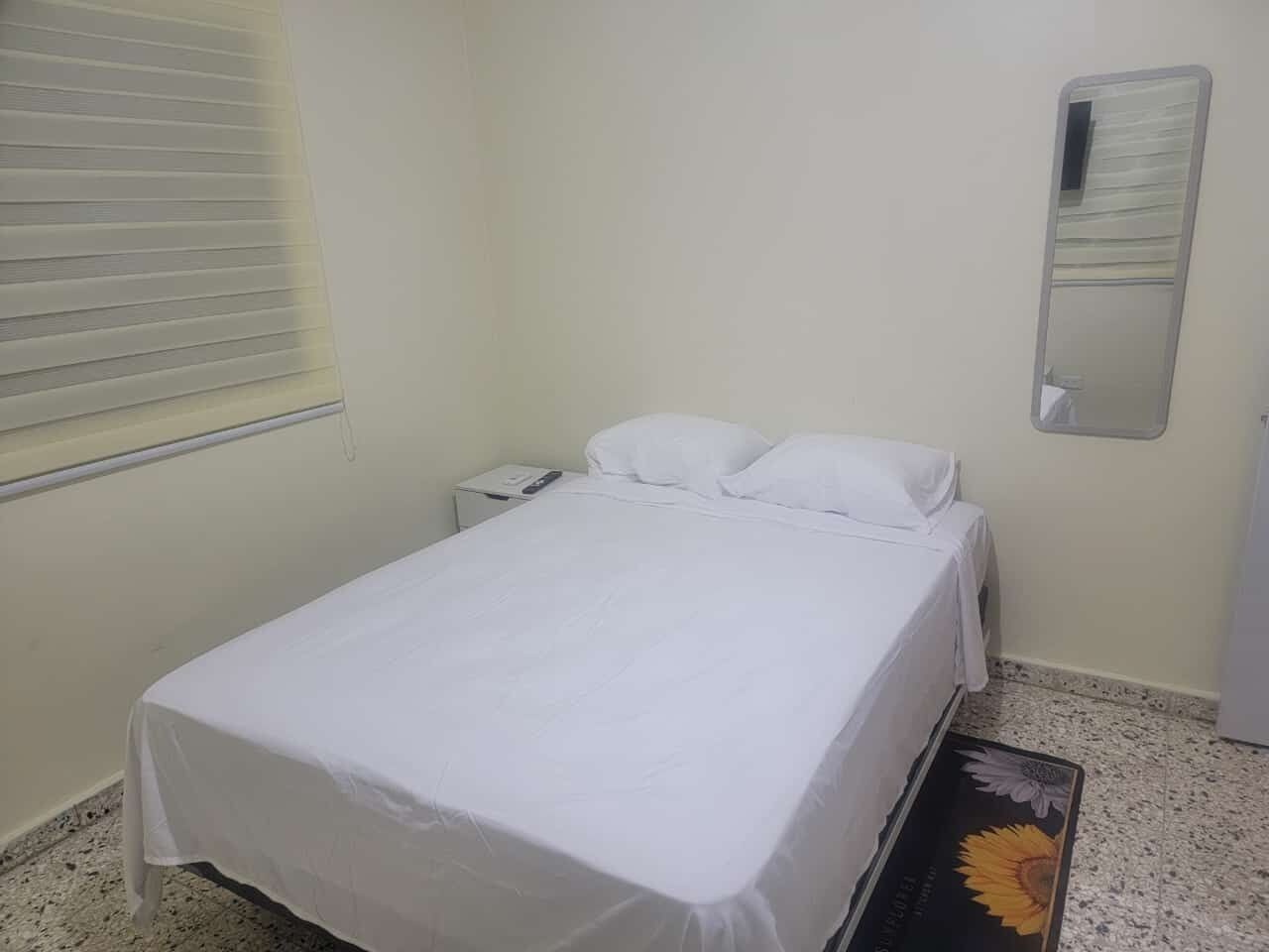 Een eenpersoonskamer met een wit sprei, een nachtkastje met lamp, een spiegel aan de muur en een raam met jaloezieën.