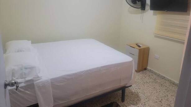 A small, clean room with a bed, a bedside drawer, an air conditioning unit, and a wall-mounted TV.