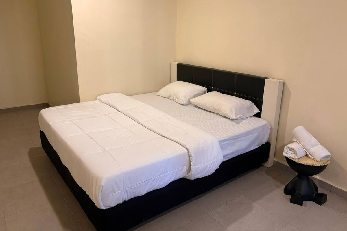 A neatly made bed with white linens in a simple room with a black headboard and a small stool with towels.