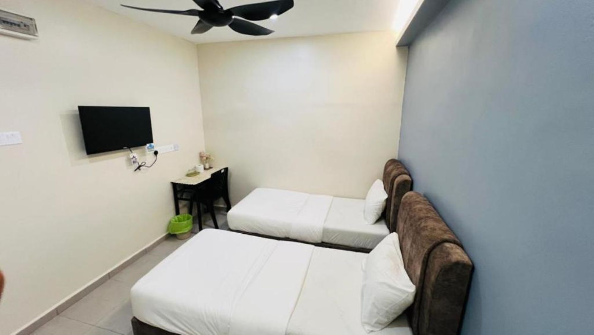 Una habitación pequeña y limpia con dos camas, un escritorio, una silla, un televisor y un ventilador de techo.