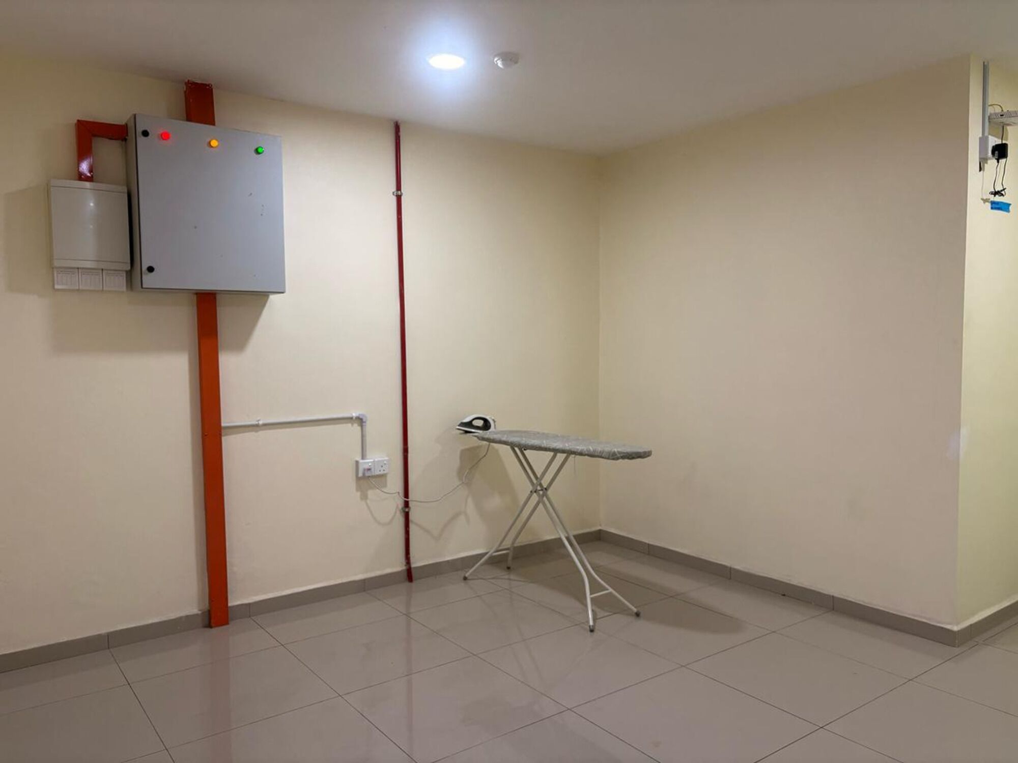 A room with a tiled floor, a wall-mounted electrical box, a red pipe, and a portable ironing board.
