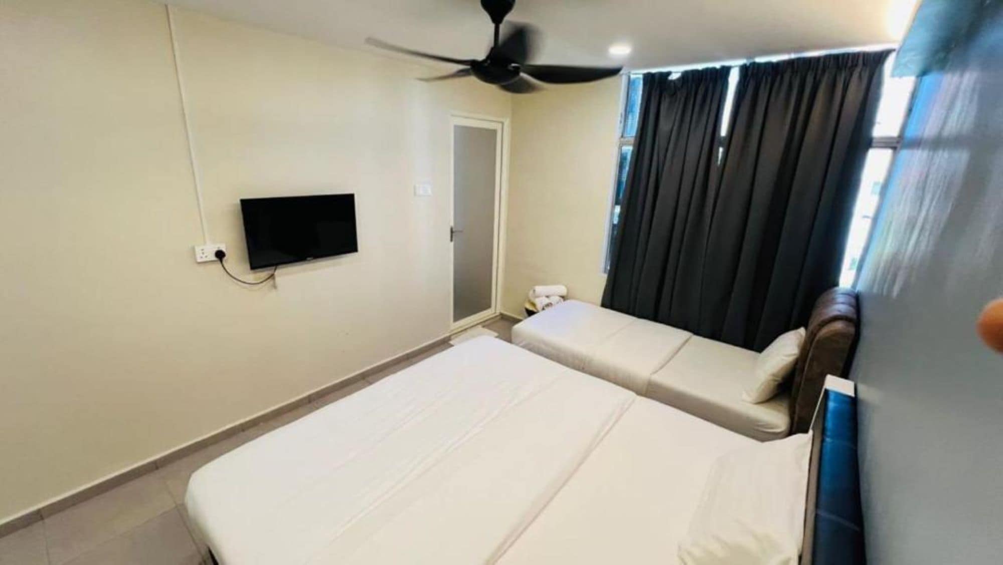 Una habitación de hotel con una cama, un televisor montado en la pared, un ventilador de techo y una ventana con cortinas.