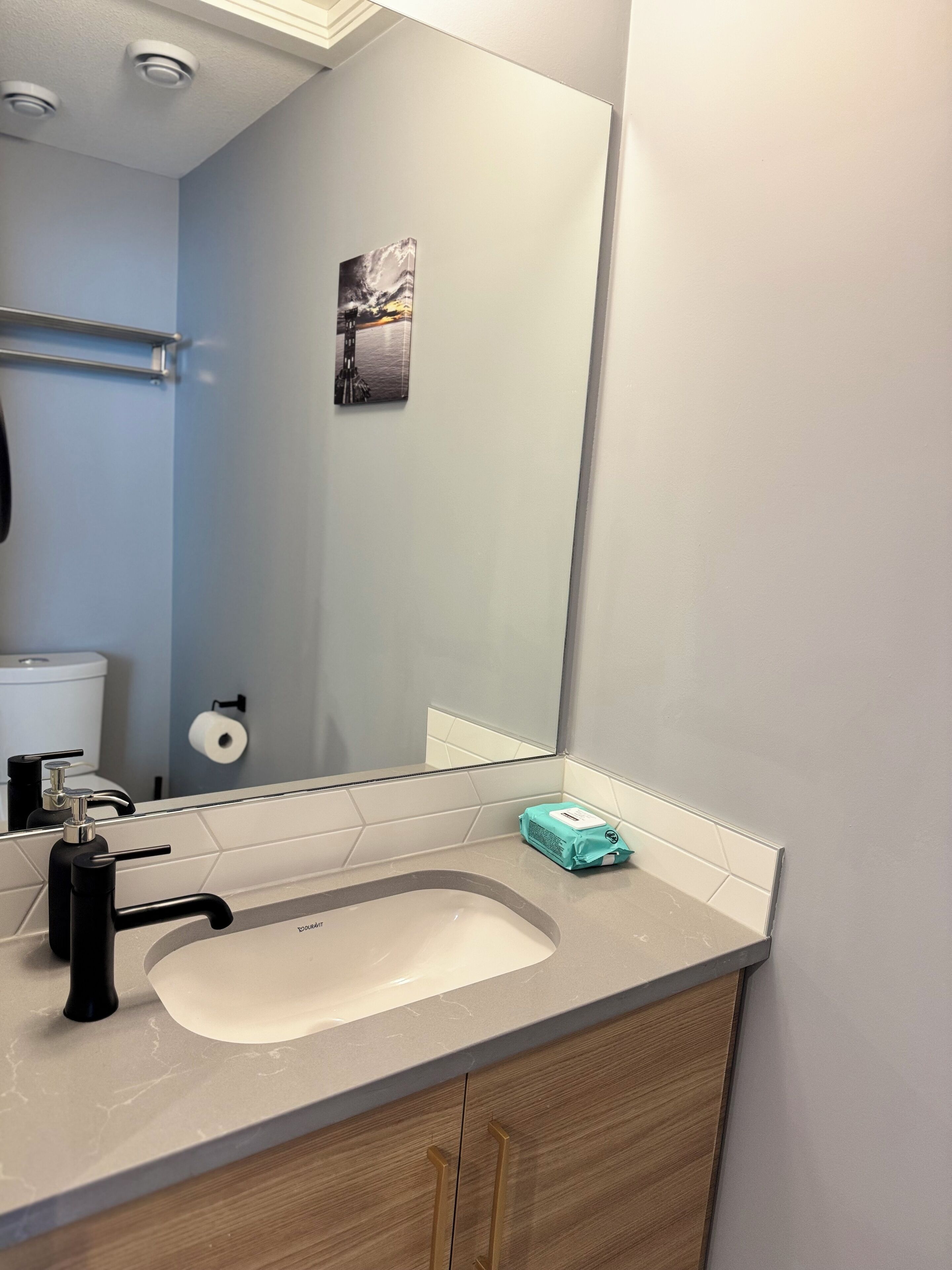 Un baño con un lavabo blanco, grifo negro y un espejo encima. Hay un dispensador de papel higiénico en la pared y un objeto azul pequeño en la encimera.