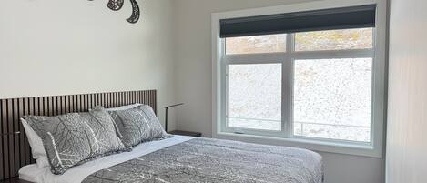 A bedroom with a bed, a nightstand, a window with blinds, and wall art.