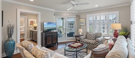 A spacious living room with a large TV, comfortable seating, a ceiling fan, and a view through a glass door to the outside.