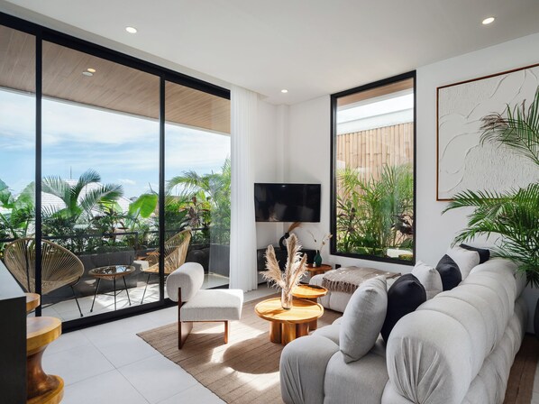 A modern living room with a large sofa, a coffee table, and a TV. There is a balcony with plants and a view of the sky.