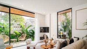 A modern living room with a sofa, coffee table, and chairs, large windows with a view of greenery, and a flat-screen TV. - A.K.A Berawa Living #2 Canggu (Canggu)