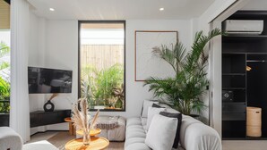 A modern living room with a sofa, coffee table, and TV. There are plants and a window with blinds. - A.K.A Berawa Living #2 Canggu (Canggu)
