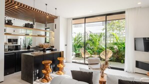 A modern kitchen with a central island, a dining area with wicker chairs, and a large sliding glass door leading to a lush garden. - A.K.A Berawa Living #2 Canggu (Canggu)