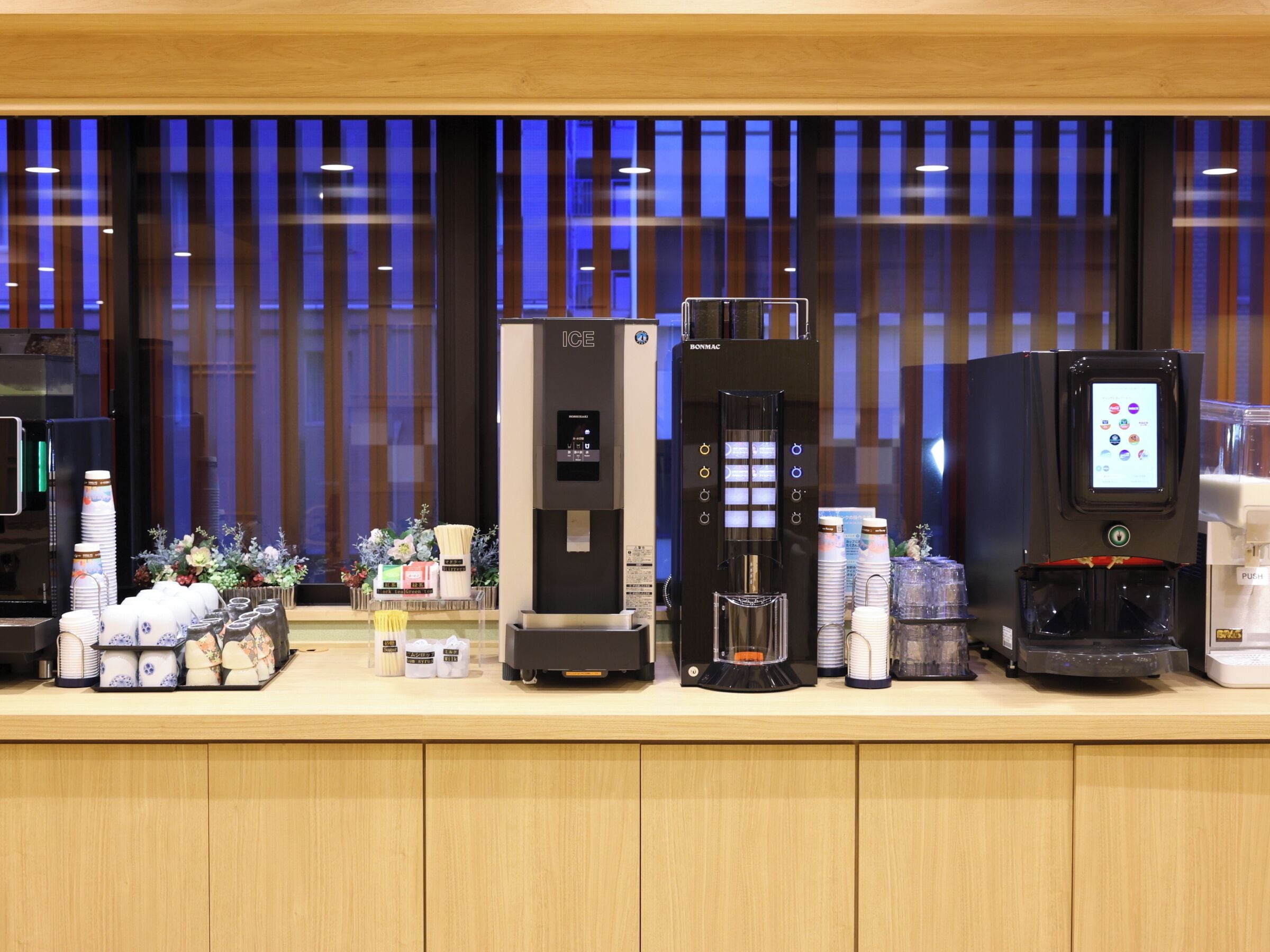 A row of beverage dispensers, including an ICE machine and a coffee maker, with various cups and containers on a counter.