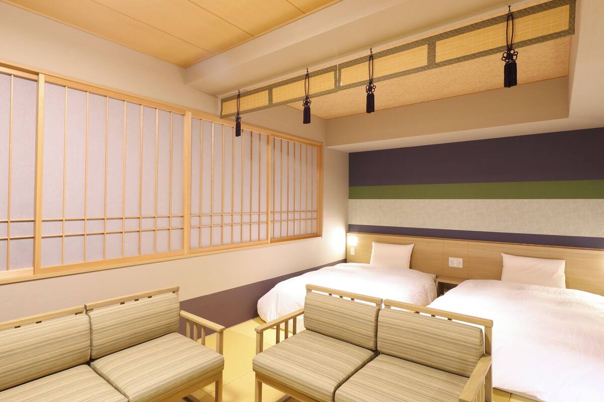 Una habitación de hotel con una pantalla tradicional japonesa shoji, una cama con ropa de cama blanca, dos mesitas de noche y un sofá.