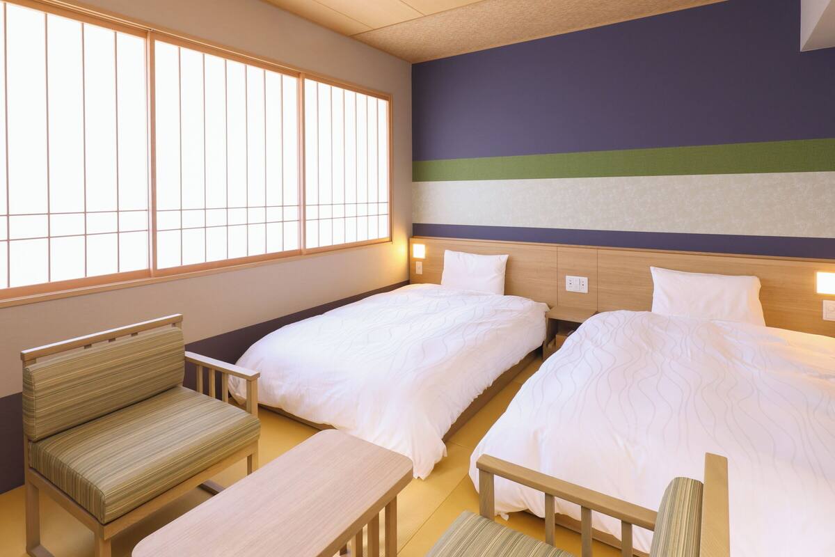 Habitación de hotel con dos camas, zona de estar y ventana con persianas verticales.