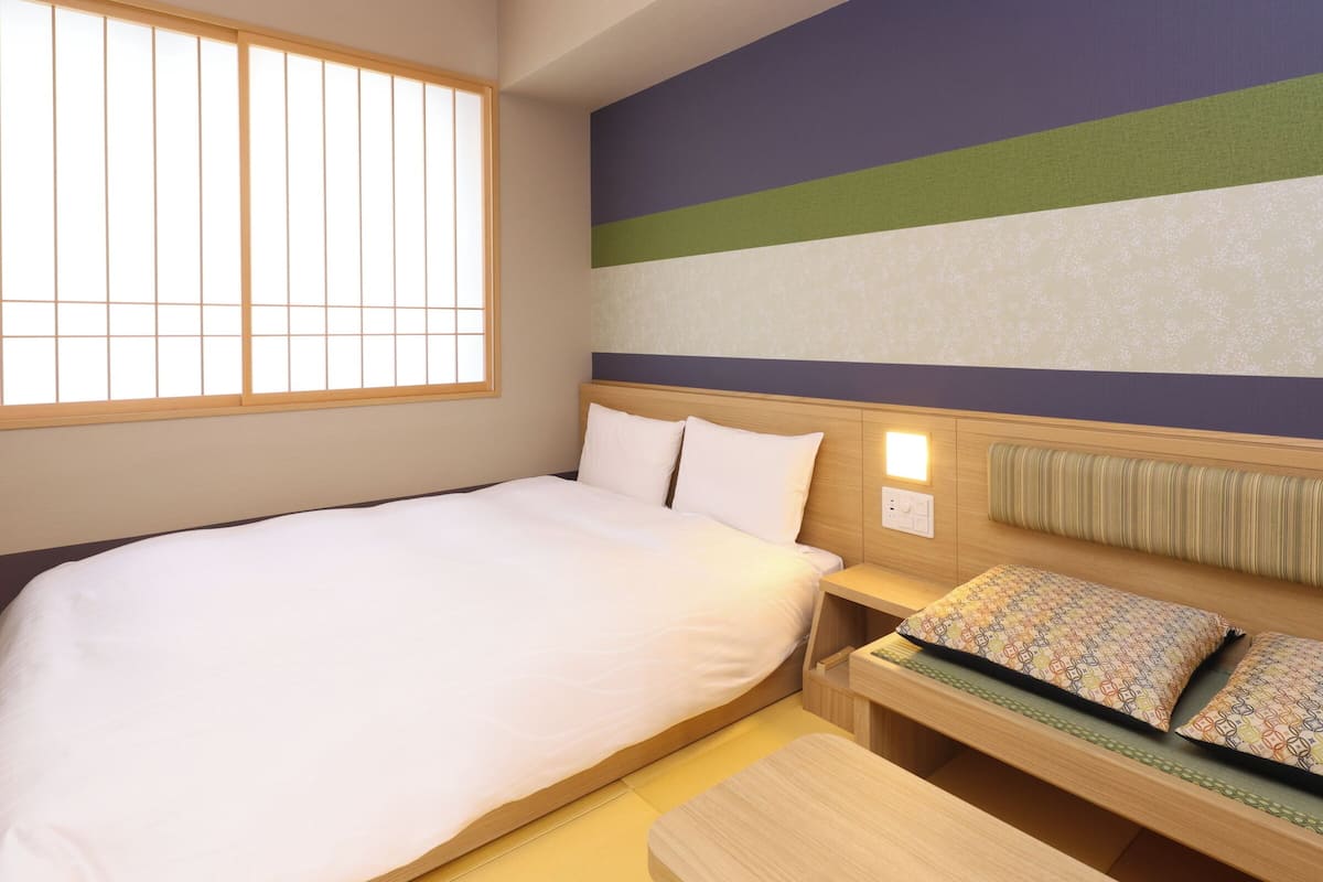 Una habitación de hotel con una cama grande, una mesita de noche con lámpara, un pequeño sofá con cojines estampados y una ventana con persianas verticales.