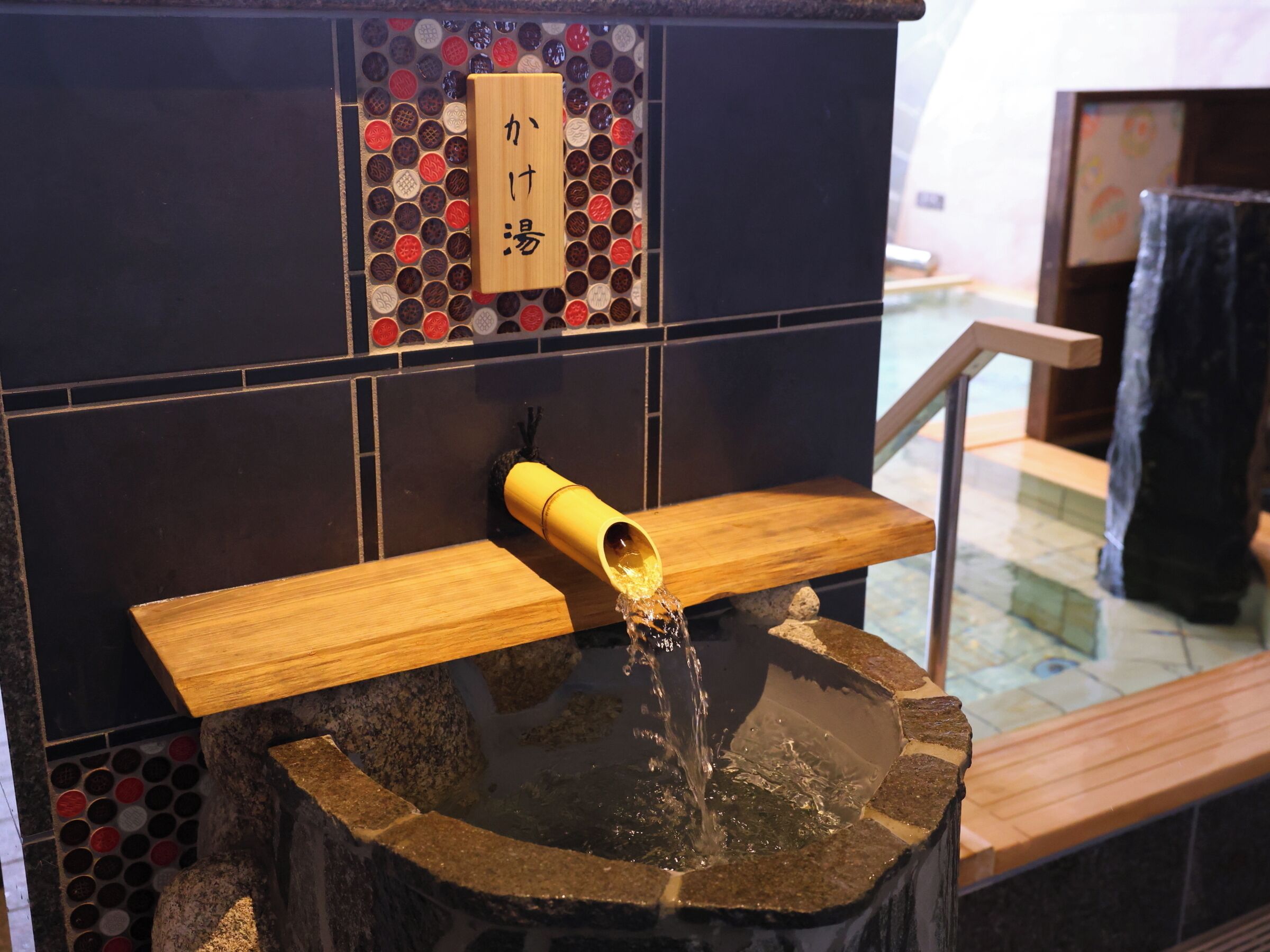 Una cuenca de piedra con agua fluyendo desde un caño de bambú, un estante de madera y una pared de azulejos con un motivo decorativo.