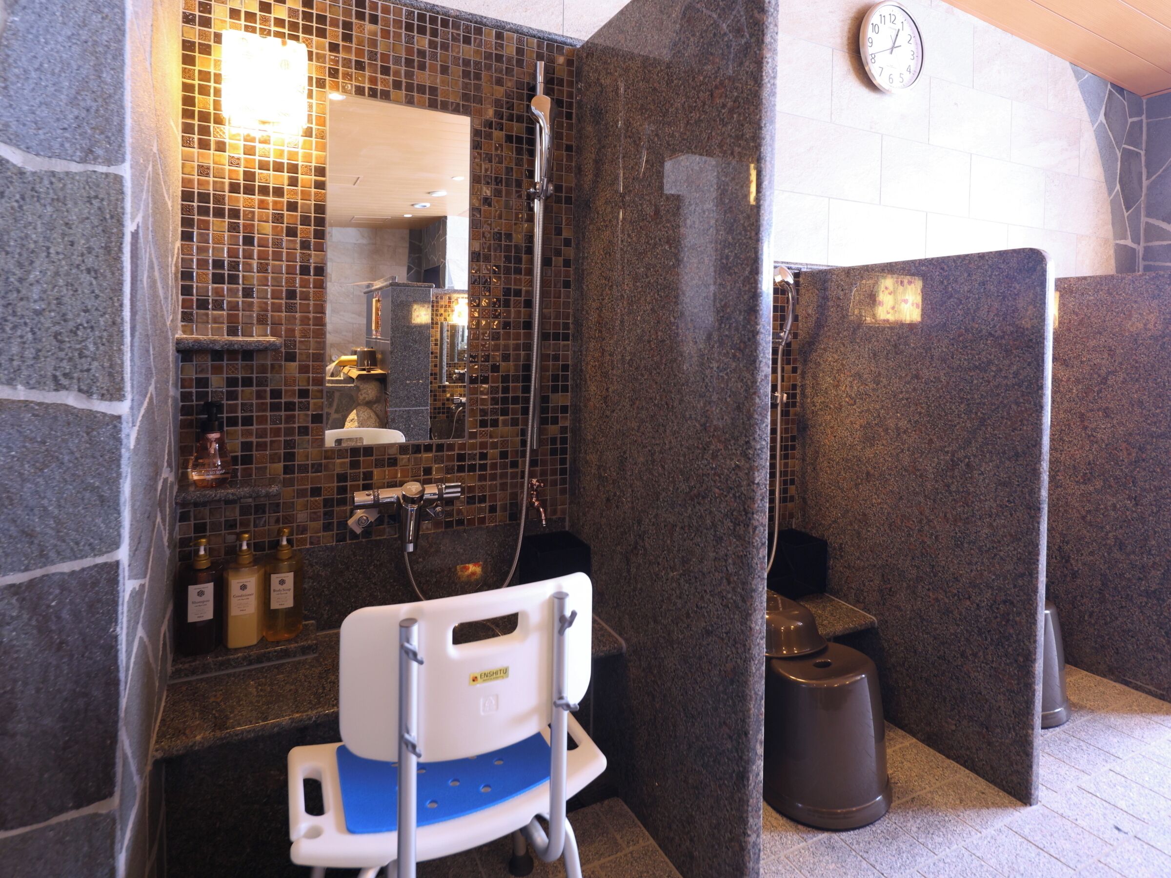 A bathroom with a shower, a sink, and a mirror. There is a wheelchair in the corner.