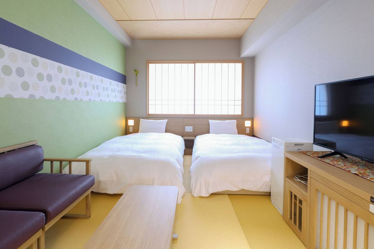Habitación de hotel con dos camas, un sofá, un televisor y una ventana que deja entrar luz natural.