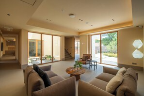 A spacious living area with a sofa set, a coffee table, and a chair. There are large windows providing natural light and a view of the outdoors.