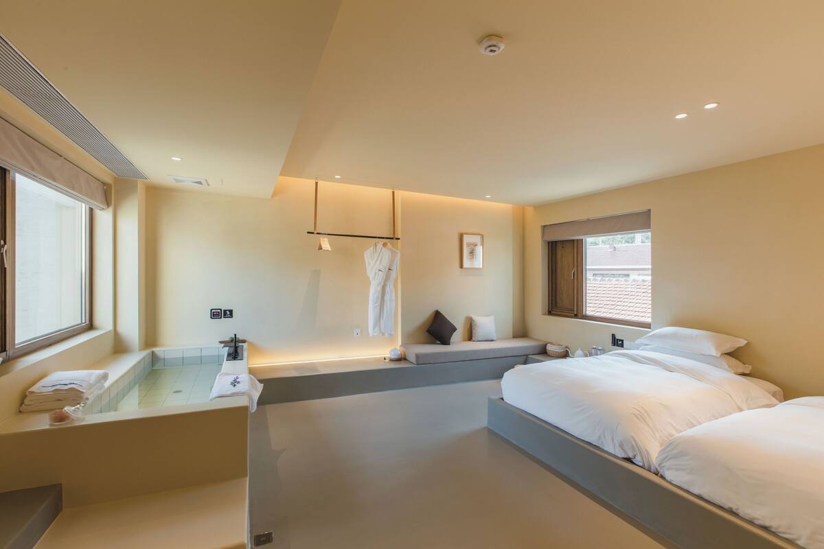 A modern hotel room with a bathtub, a seating area with a sofa and pillows, a large bed, and a window providing natural light.