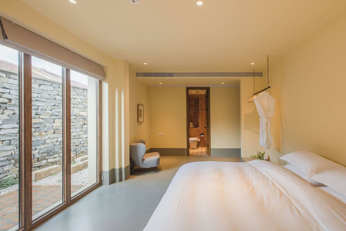 A modern hotel room with a large bed, a seating area, and a view of a stone wall outside.