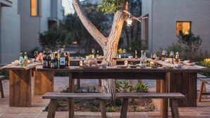 Mobiliario para comedor al aire libre con varias mesas y bancos de madera, dispuestos alrededor de un árbol central.