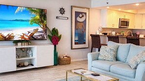 Een woonkamer met een lichtblauwe bank, een salontafel, een tv waarop een strandafbeelding wordt getoond, een keuken op de achtergrond en een decoratief kunstwerk aan de muur.