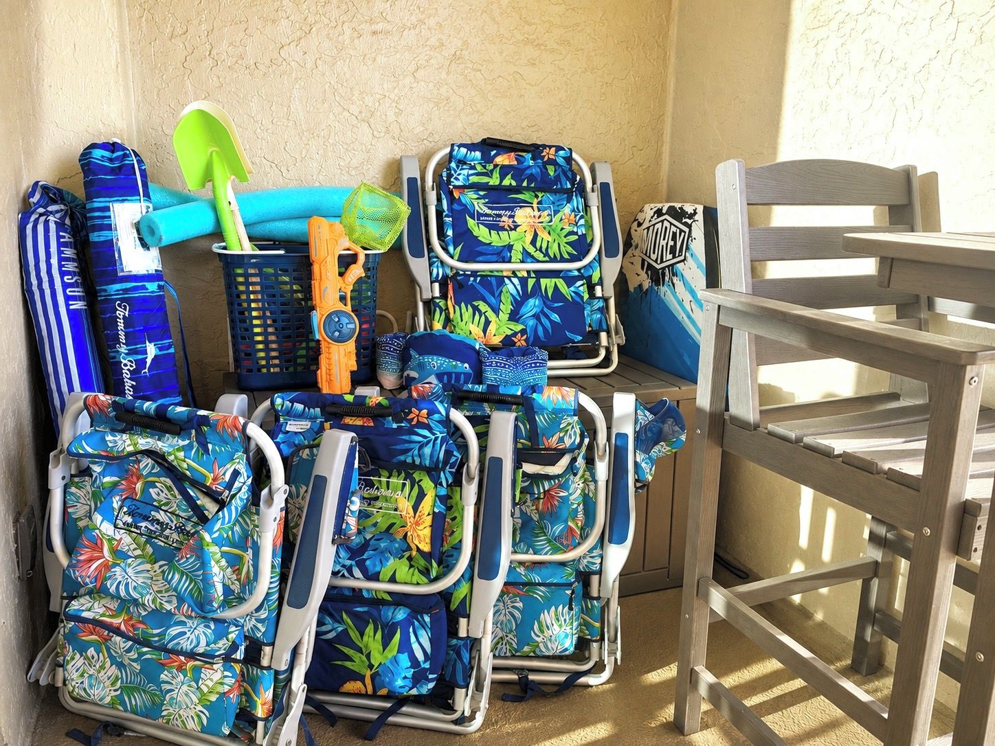 A corner of a room with a wooden chair, a metal table, and several colorful backpacks and a beach bag.