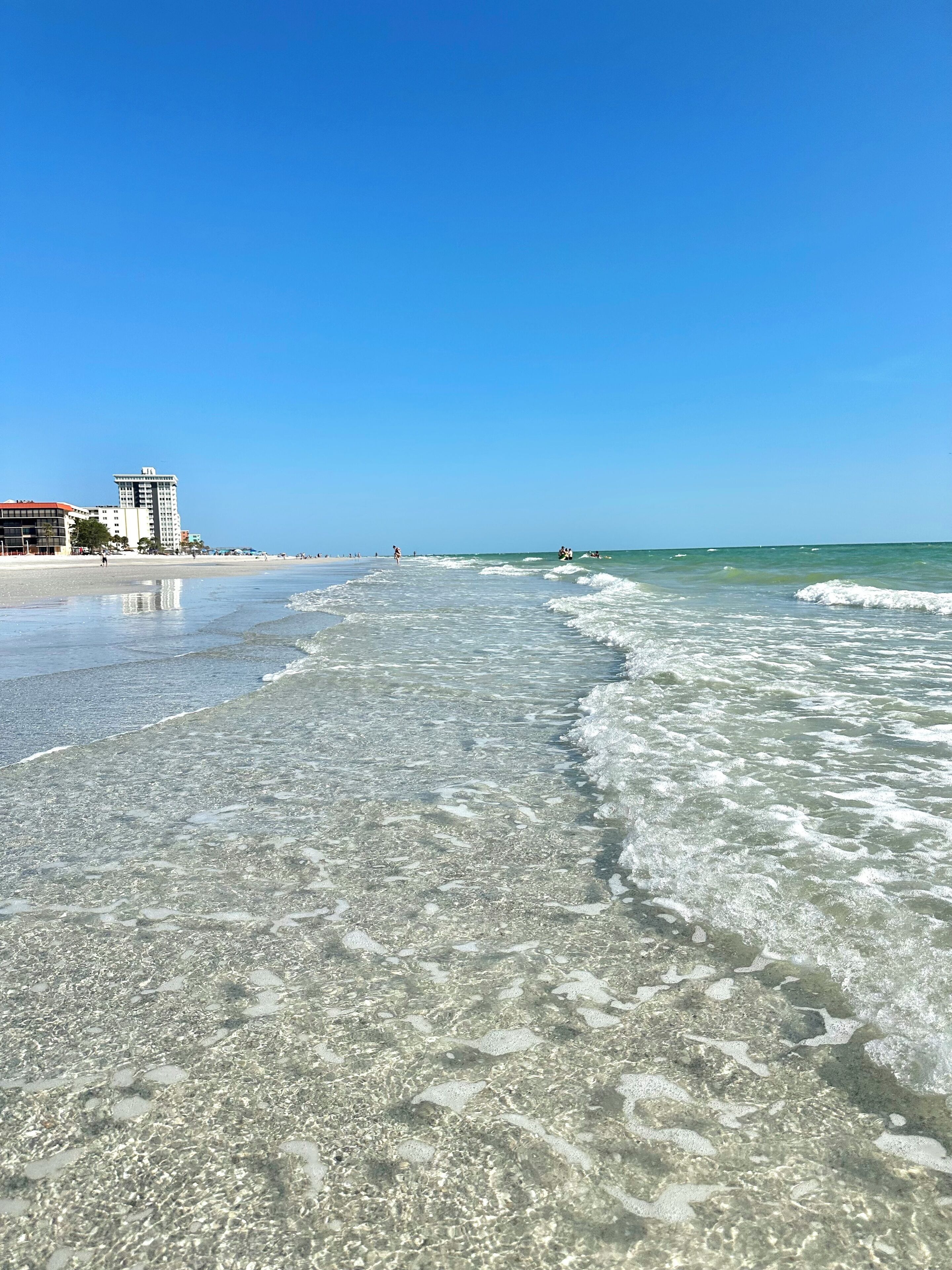 A beach with clear shallow waters, a few buildings in the background, and a clear blue sky.