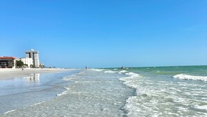 Een strand met helder ondiep water, een paar gebouwen op de achtergrond en een strakblauwe lucht.