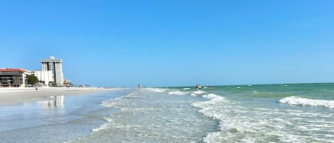 A beach with clear shallow waters, a few buildings in the background, and a clear blue sky.