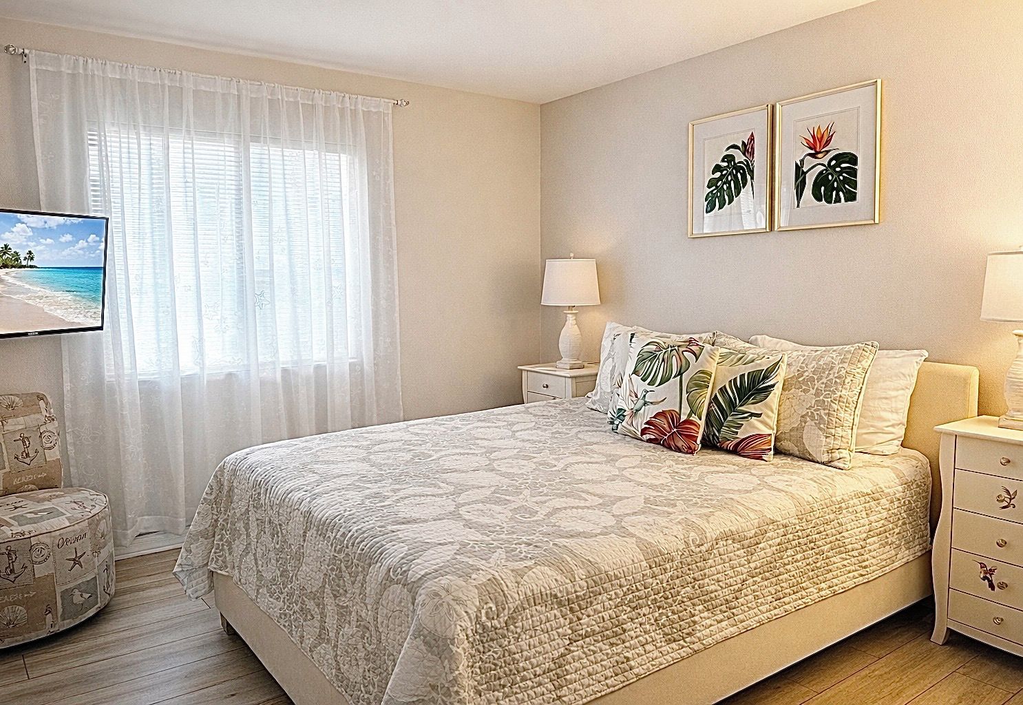 A bedroom with a large bed, a TV, a nightstand with a lamp, and tropical-themed wall art.