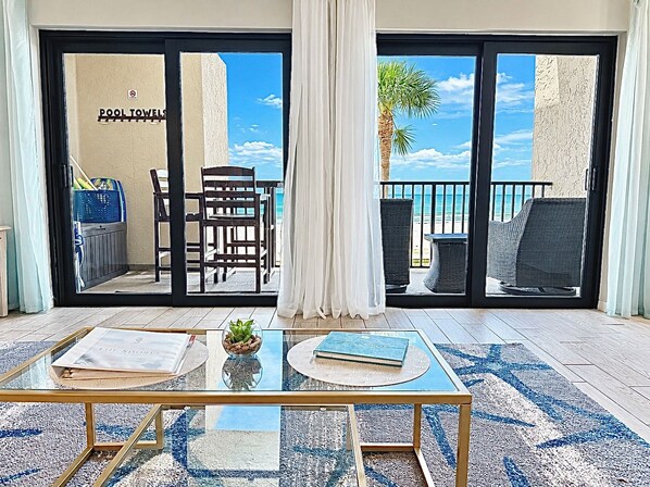 A room with a glass table, a chair, a small plant, and a view of the beach through a sliding door.