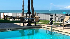 A pool area with clear blue water, surrounded by lounge chairs and a table, with a view of the ocean and a fence in the background. - Beachfront 2BR Condo. Ocean view. Quiet Small Building. No Crowds. Heated Pool. (Redington Shores)