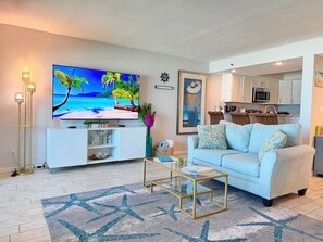 A living room with a flat-screen TV, a sofa, a coffee table, and a kitchen in the background.