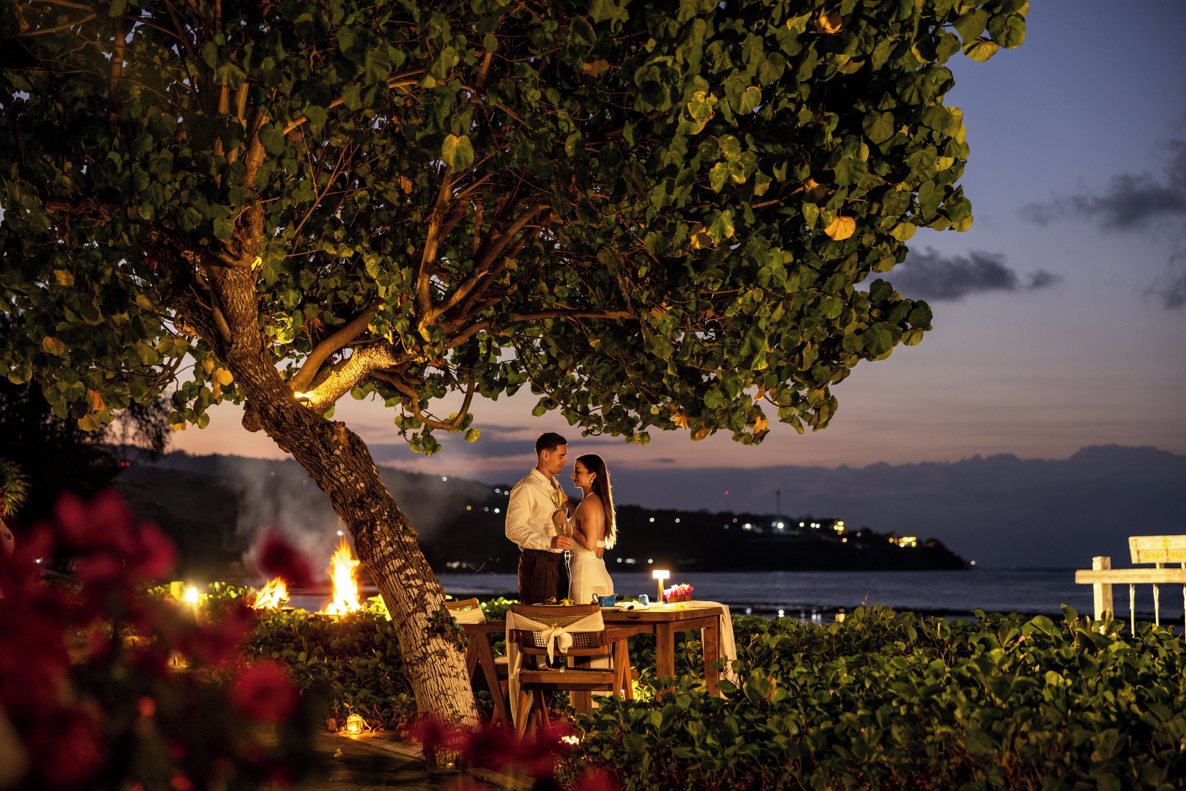A couple standing under a tree with a table and chairs, overlooking a body of water.