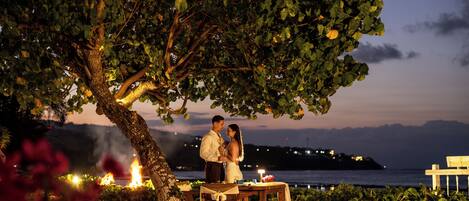 A couple standing under a tree with a table and chairs, overlooking a body of water.