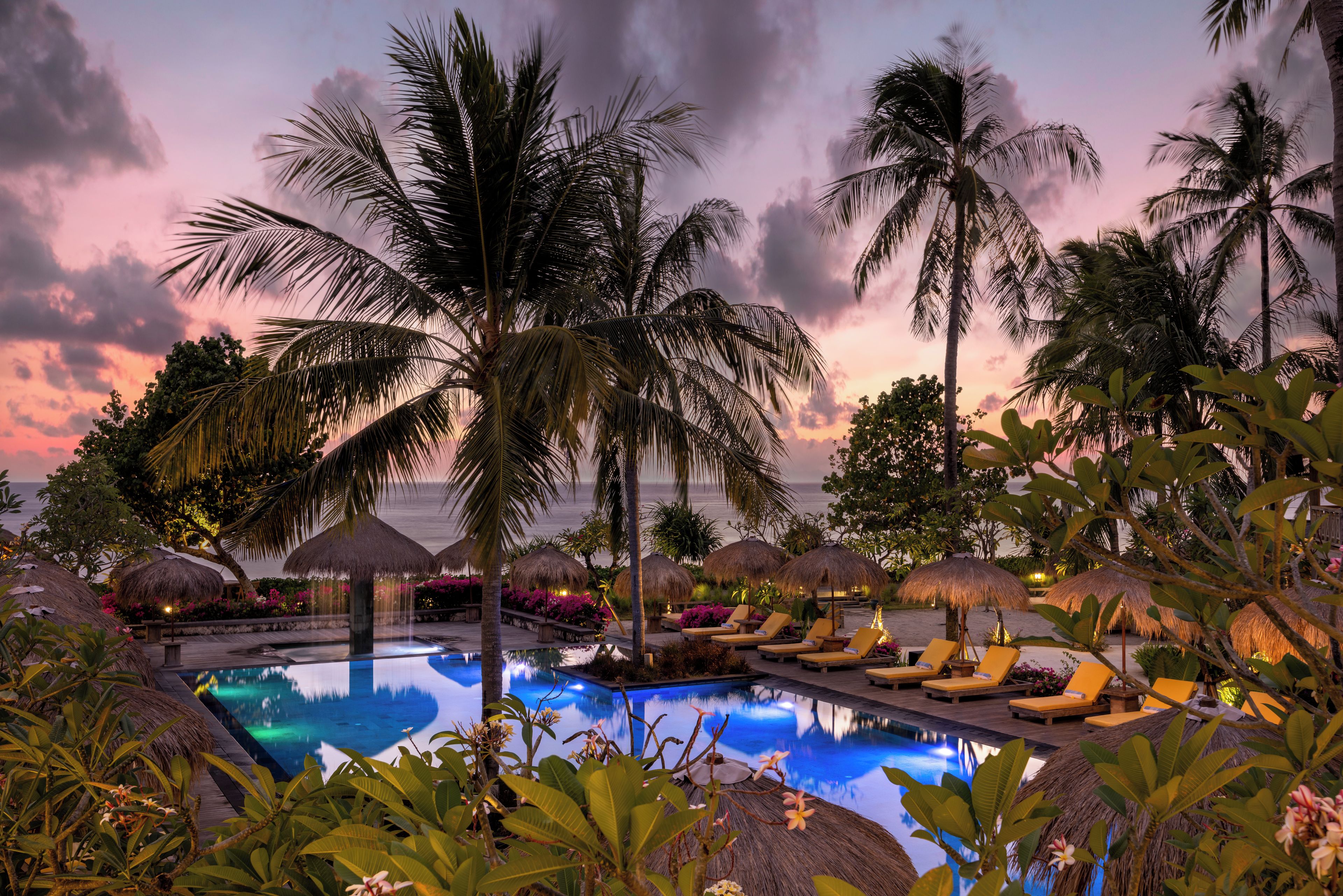 A pool area with lounge chairs and thatched umbrellas, surrounded by palm trees and tropical plants.