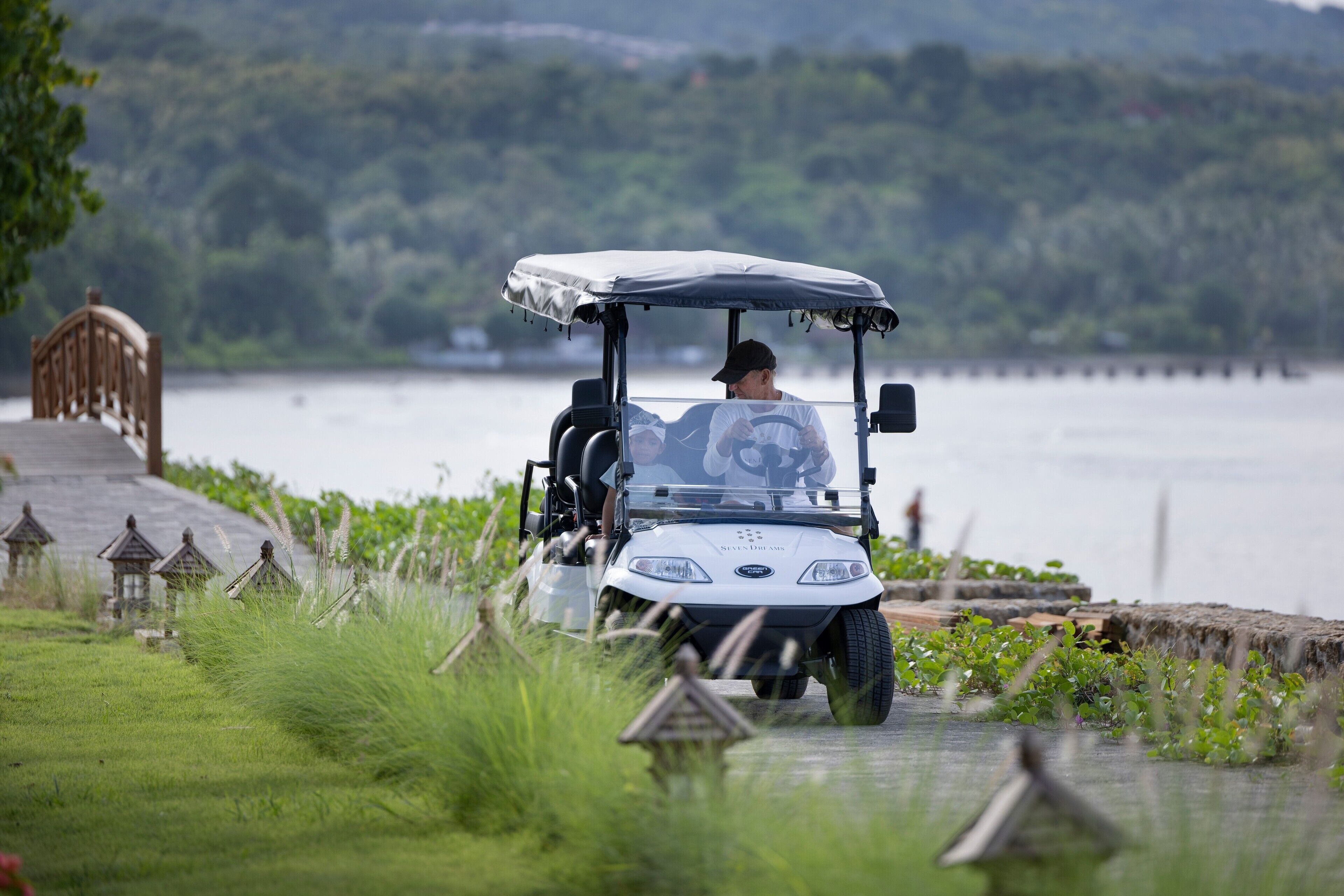 A golf cart on a grassy path near a body of water with a forested background.