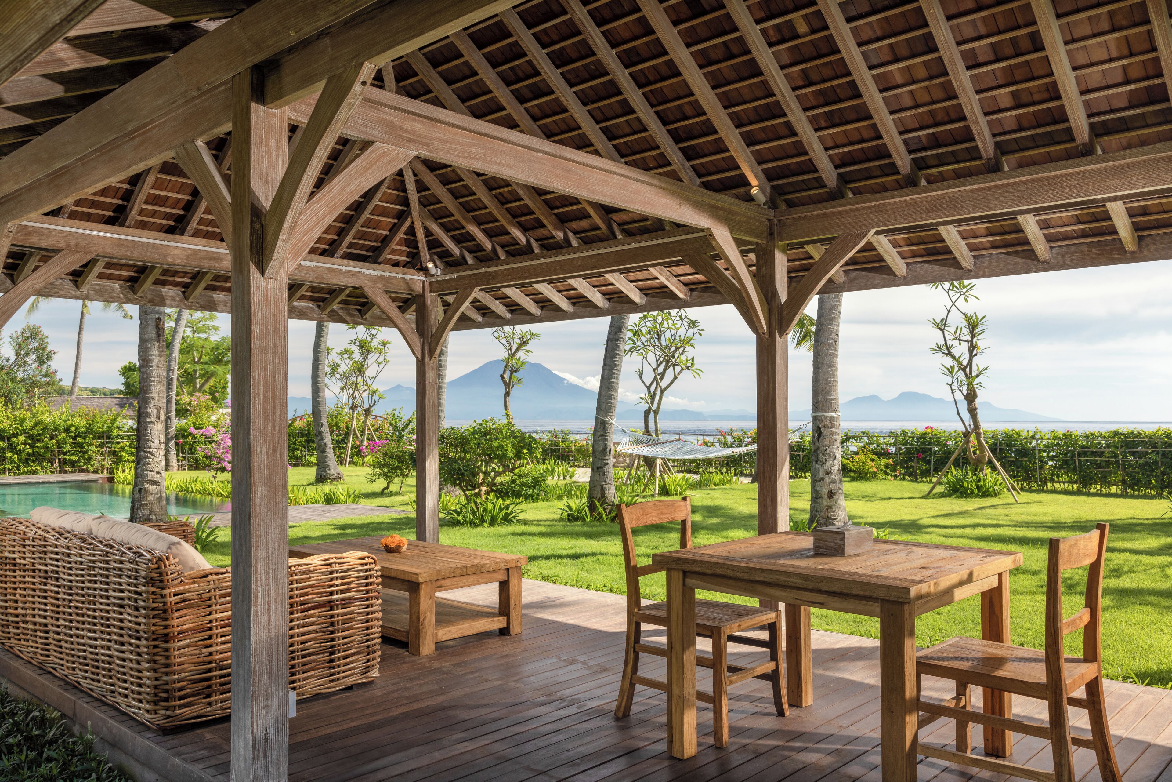 Eine Holzterrasse mit einer Pergola, Gartenmöbeln und Blick auf ein Schwimmbad und Berge.