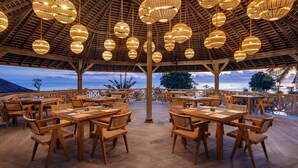 An outdoor dining area with wooden tables and chairs, surrounded by a wooden fence, and hanging wicker lamps. - Seven Dreams A Pramana Experience (Penida Island)