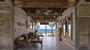 A wooden interior with a sign reading "SEVEN DREAMS SPA," a ceiling fan, wicker lamps, a bench, and a view of the ocean. - Seven Dreams A Pramana Experience (Penida Island)