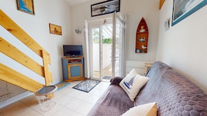 A room with a wooden staircase, a television on a cabinet, a sofa with cushions, a small table, and a door leading outside. - Le Lagon Bleu (Bretignolles-sur-Mer)