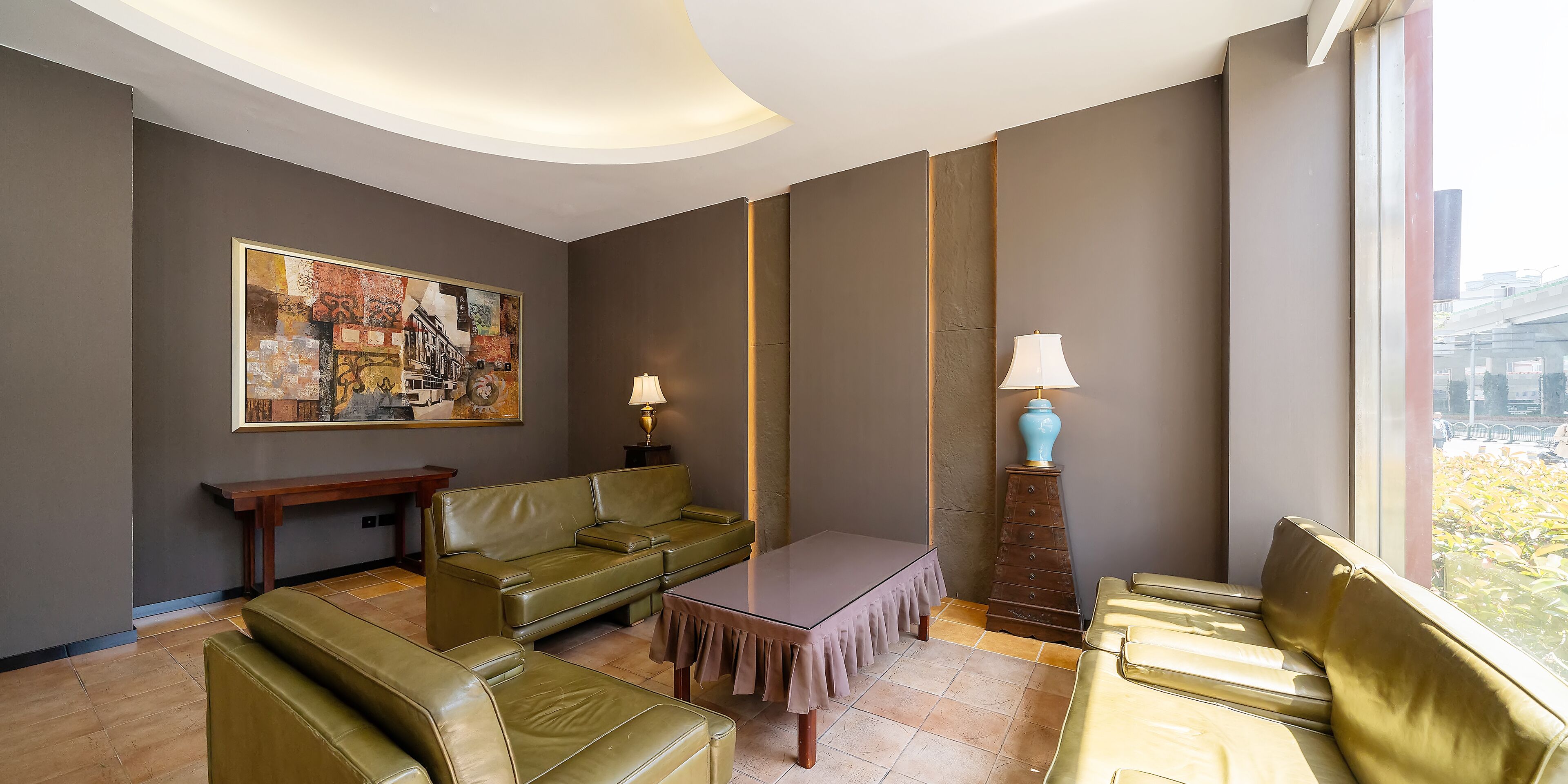 A modern hotel lobby with a green leather sofa, a wooden coffee table, and a dining table with a white tablecloth. There is a large painting on the wall and a lamp on a wooden stand.