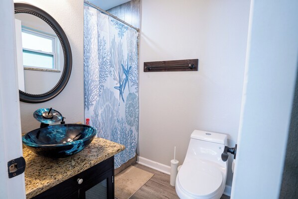 A bathroom with a round mirror, a blue glass bowl sink, a toilet, and a shower curtain with a leaf pattern.