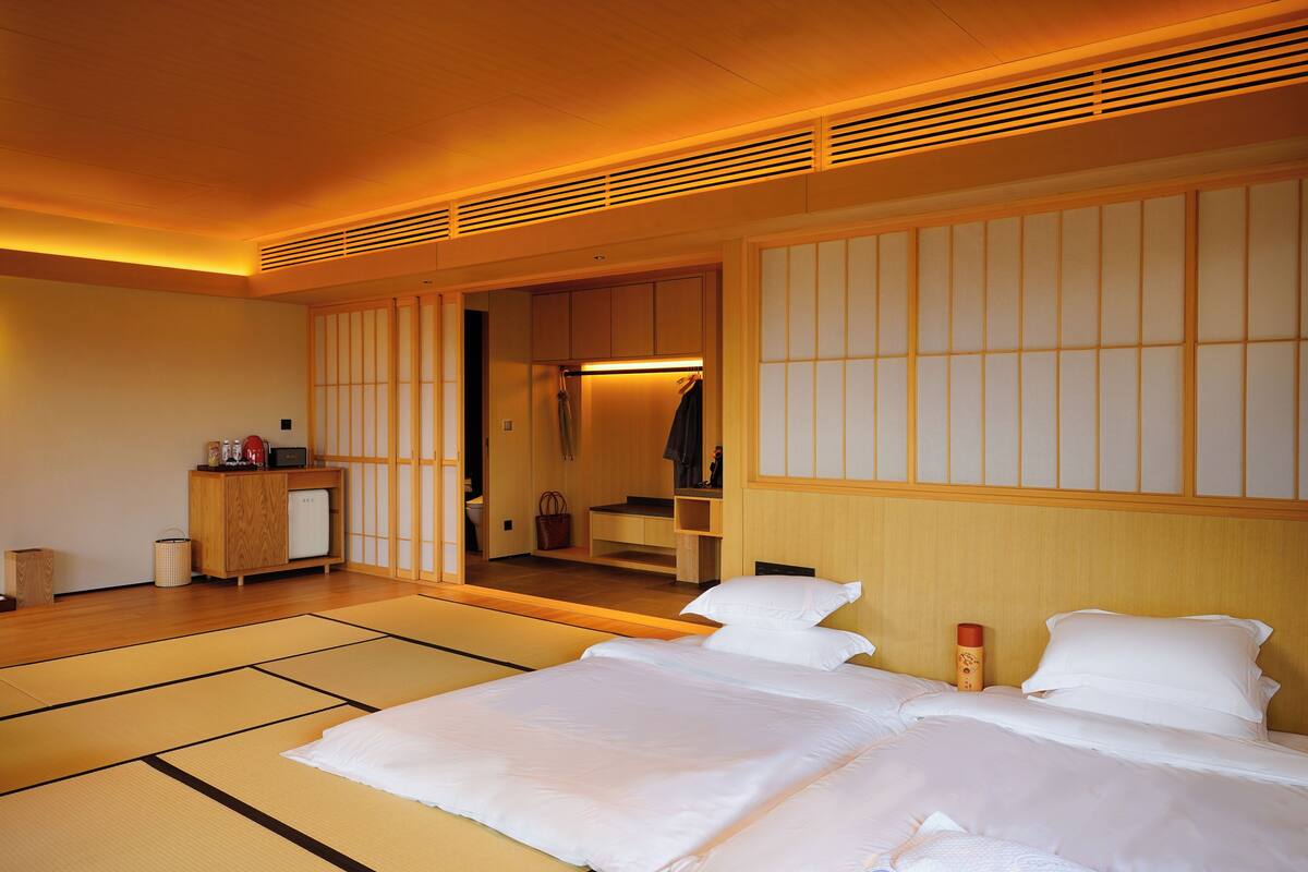 Un dormitorio de estilo japonés tradicional con suelo de tatami, una cama con ropa de cama blanca, un armario de madera y una puerta corredera que da a un baño.