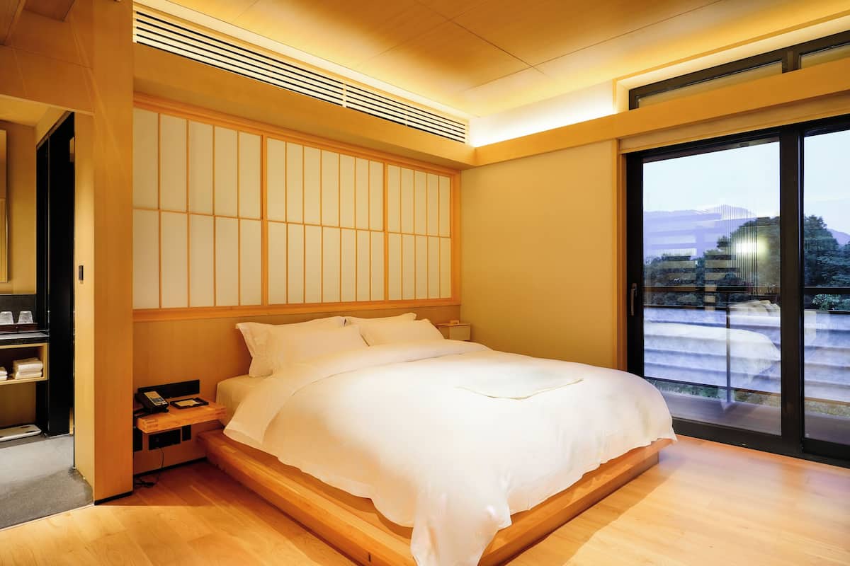 Una habitación de hotel moderna con una cama de madera, un armario empotrado y un amplio ventanal corredero que da a un balcón con vistas a la ciudad.