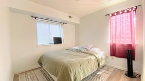 Een slaapkamer met een bed, een raam met zonwering, een staande ventilator en een kamerbreed tapijt met een patroon.