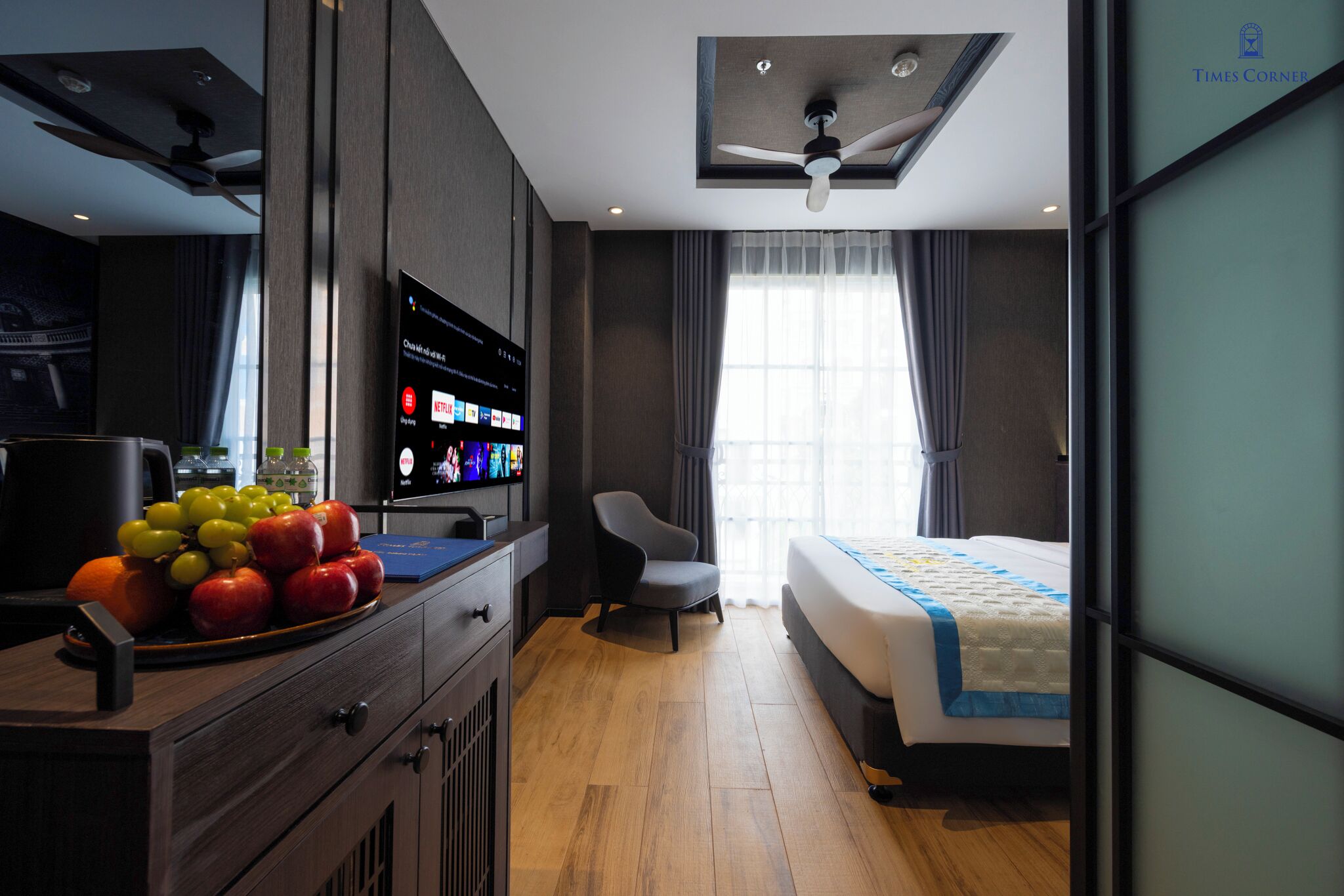 A modern hotel room with a bed, a desk with a fruit bowl, a television, and a ceiling fan.
