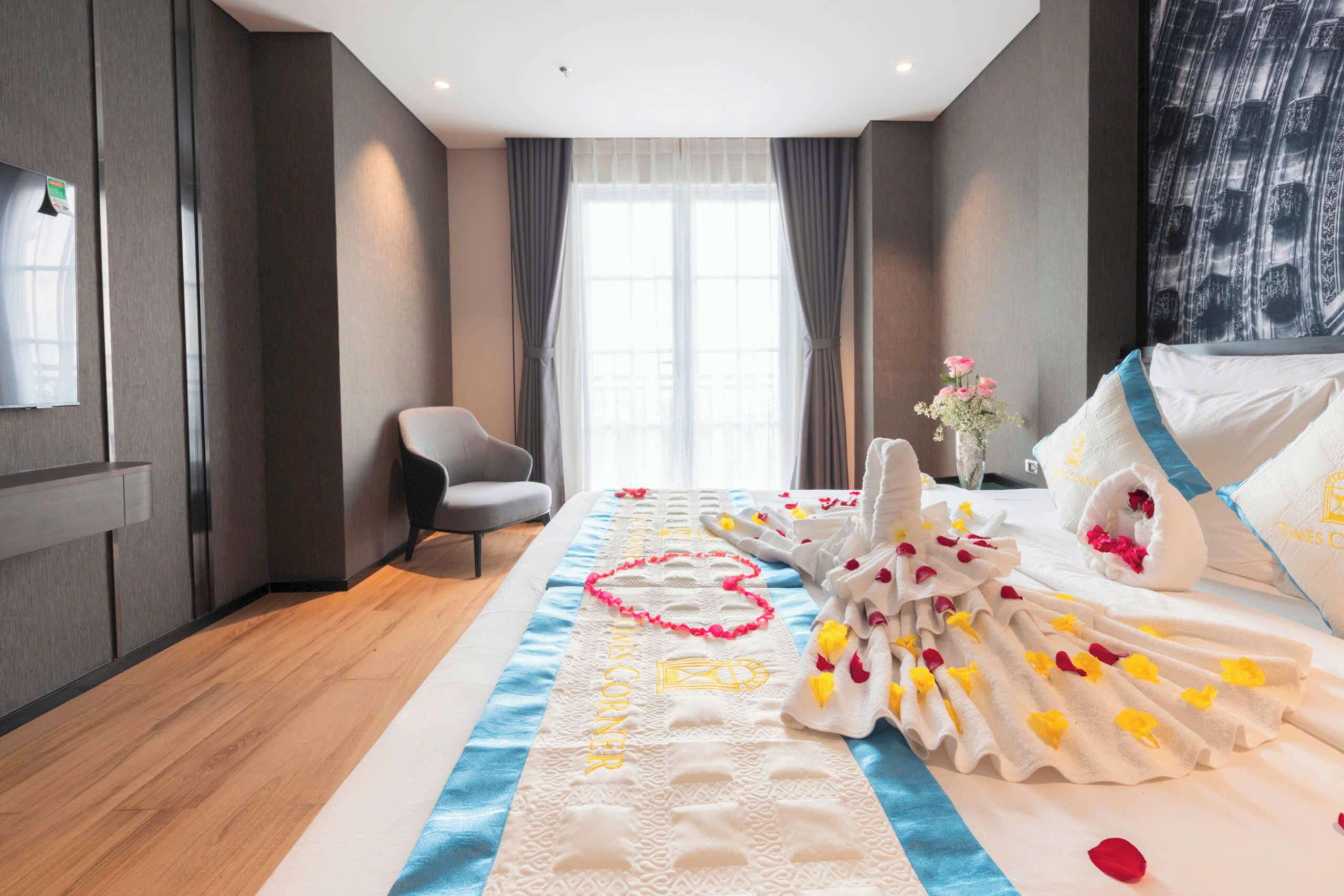 A hotel room with a bed adorned with a heart-shaped towel arrangement, a blue and white striped runner, and a vase with pink flowers.